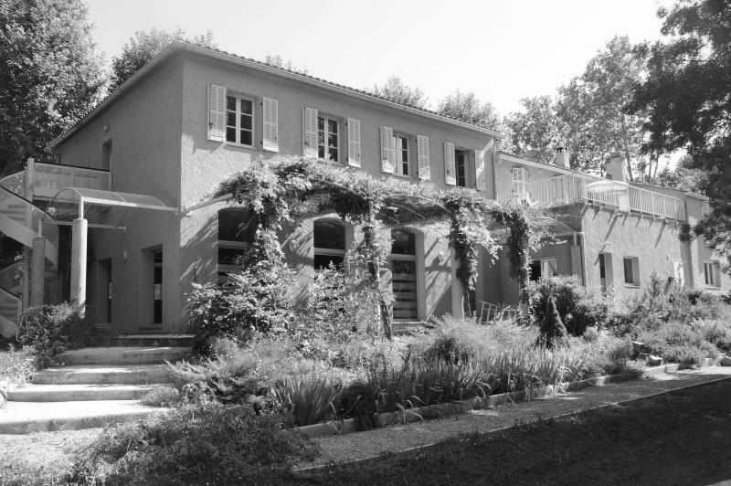 Black-and-white photo of the Shumeikan at Bras: a two-story Provençal building with shuttered windows, a vine-covered pergola along the ground floor, stone steps and a planted garden in front, with tall trees behind.