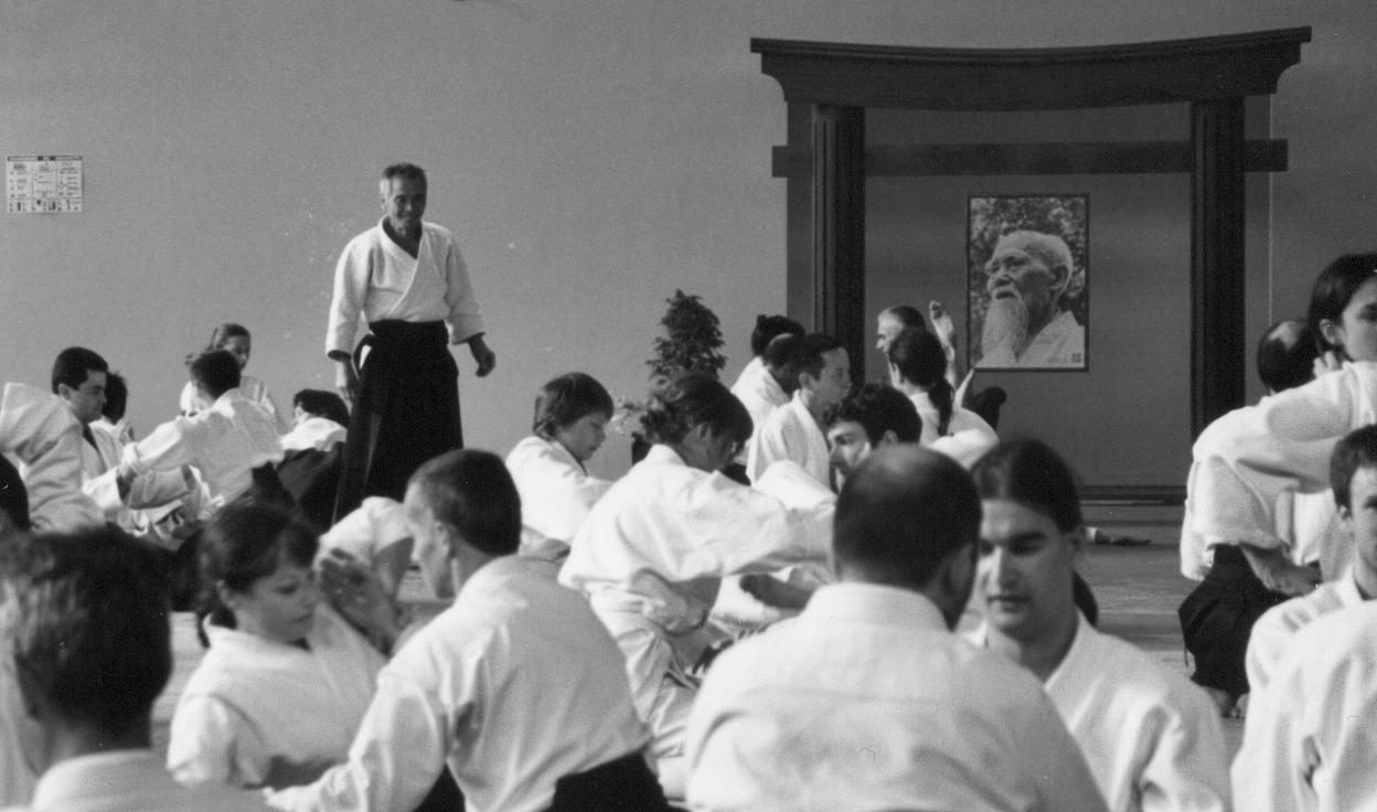 Black-and-white photo: Tamura Shihan, in white keikogi and dark hakama, stands watching rows of students practicing in pairs on the mat at Lesneven; a portrait of O-Sensei hangs on the dojo wall behind them.